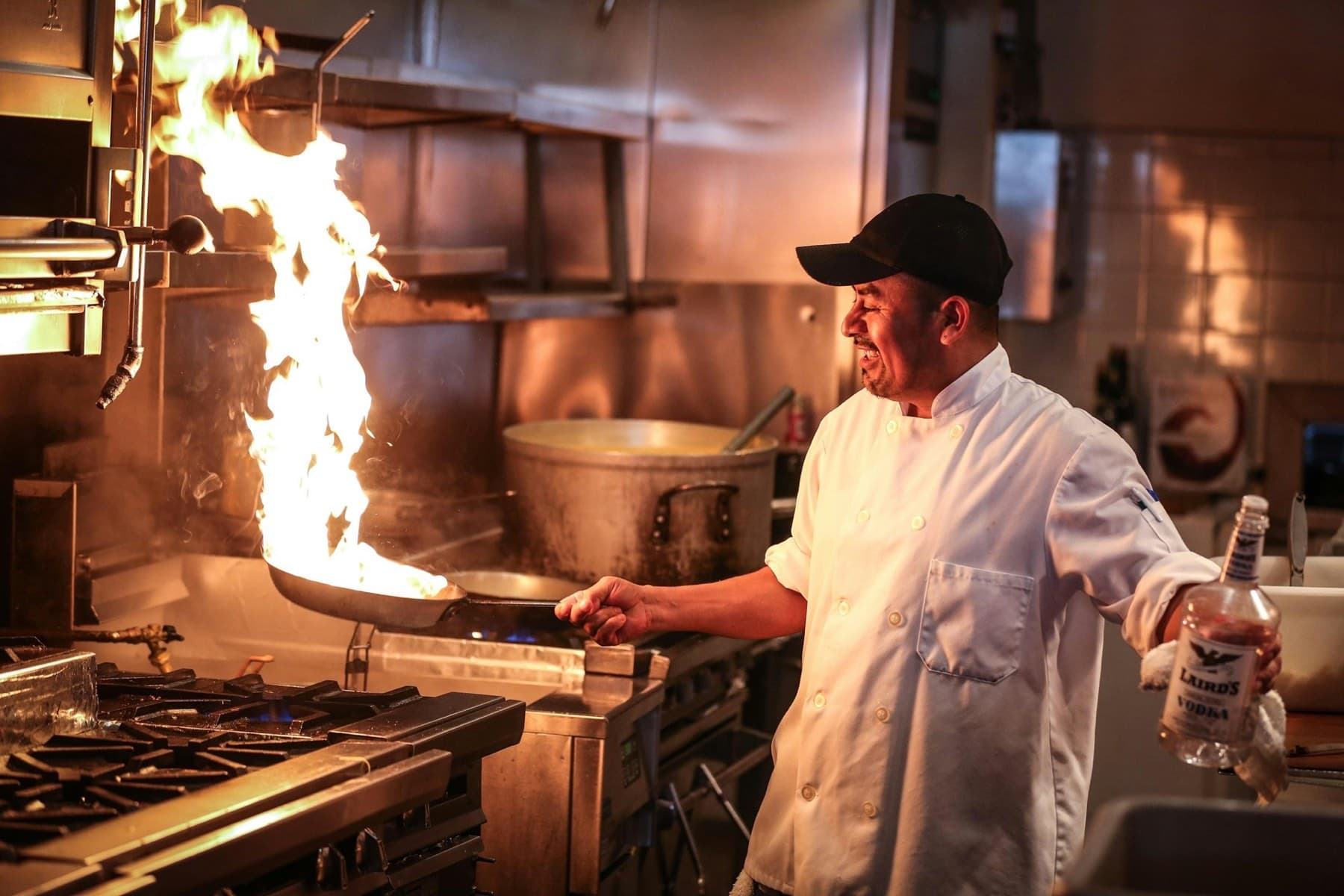 A cook in chef's whites flambéing a pan on a busy restaurant range